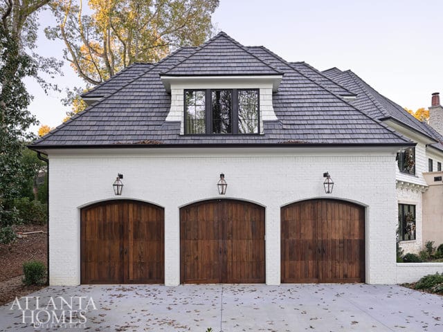 White brick garage with three arched wooden doors, black dormer window, and dark shingle roof.