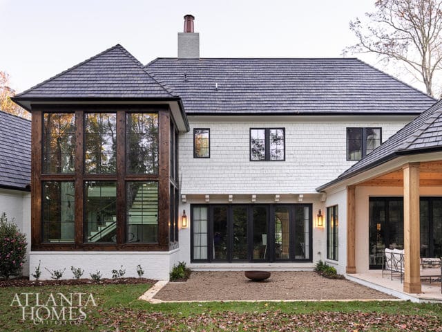 Modern home exterior with white shingle siding, dark-framed windows, and a multi-level dark shingle roof. A large glass-walled section reveals an interior staircase.