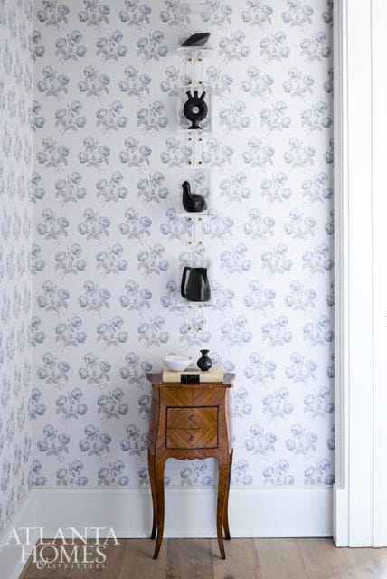 Hallway with blue floral wallpaper, a wooden accent table with drawers, and clear acrylic shelves displaying black decorative objects.