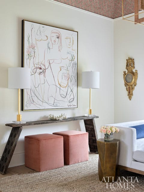 Living room with abstract line art above a rustic console table, two blush pink ottomans, and a white sofa with a navy pillow.