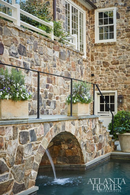 Stone house exterior with arched bridge over a pool, white-trimmed windows, and planters with flowers.