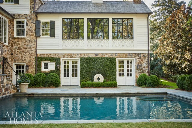 Stone and white clapboard house with a swimming pool in front. Features include French doors, manicured hedges, and large windows reflecting trees.