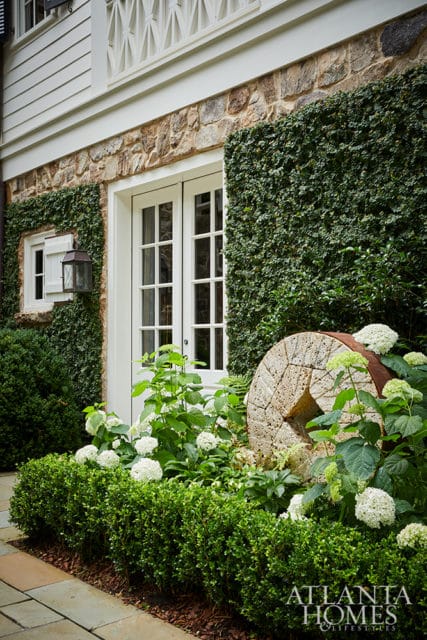 Stone and white-sided house exterior with ivy-covered walls, double glass doors, and a garden featuring hydrangeas and a millstone.