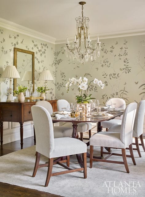 Elegant dining room with a light green floral wallpaper, a crystal chandelier, a dark wood dining table set for six, upholstered chairs, and a vintage sideboard with lamps and a mirror.