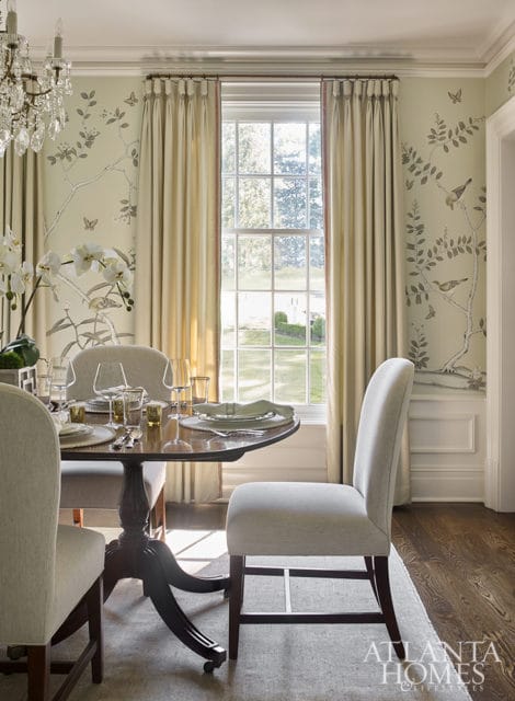 Dining room with a table set for a meal, upholstered chairs, and wallpaper featuring birds and branches. A crystal chandelier hangs overhead.