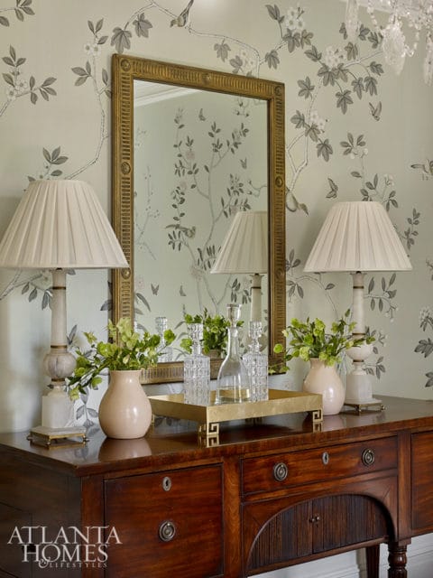 A wooden sideboard topped with two lamps, a vase of greenery, crystal decanters on a gold tray, and a gold-framed mirror reflecting chinoiserie wallpaper.