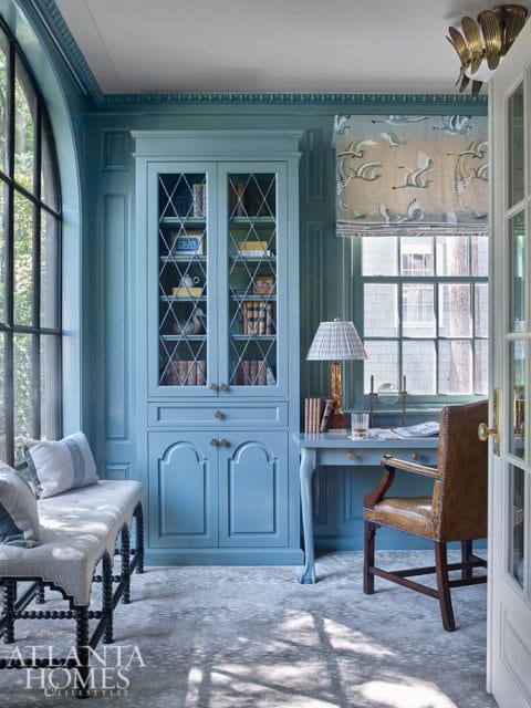 Blue study with built-in bookcase, leather armchair at desk, and window seat. Features paneled walls, diamond-patterned glass doors on bookcase, and a bird-print roman shade.