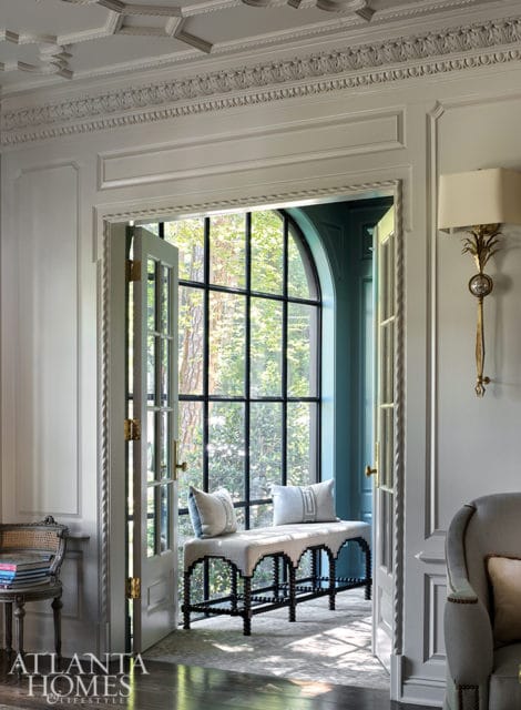 Light gray room with ornate ceiling molding, a tall arched window with black mullions, and a light upholstered bench with two pillows.