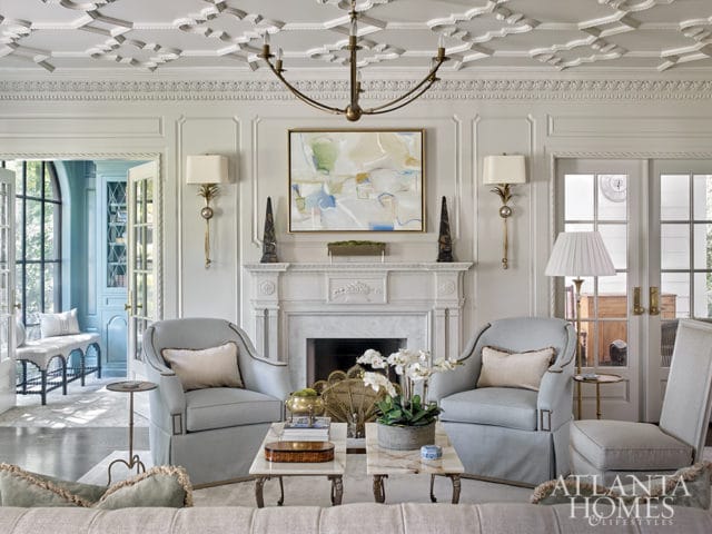 Light blue upholstered armchairs flank a marble fireplace with a contemporary abstract painting above. Intricate coffered ceiling and ornate wall molding.