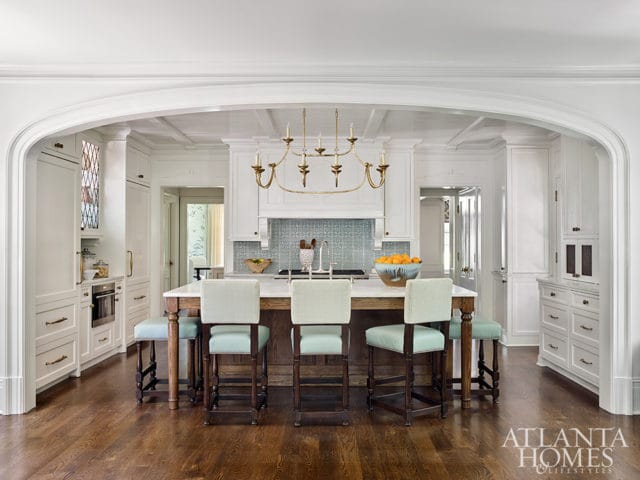 White kitchen with coffered ceiling, marble island with wooden base, light blue and white checkered bar stools, and a gold chandelier.