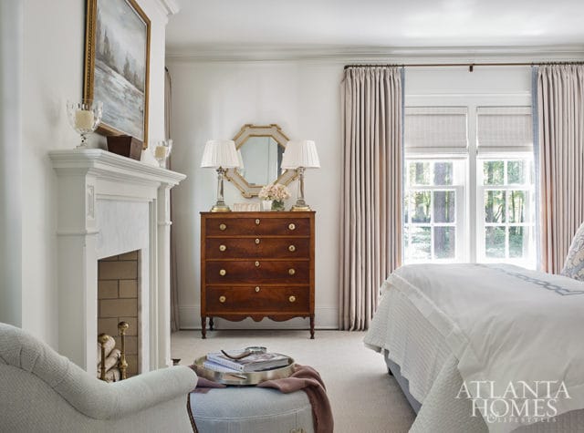 Bedroom with a white fireplace, wood chest of drawers with two lamps, and a bed with white linens. A logo for Atlanta Homes & Lifestyles is visible in the lower right corner.