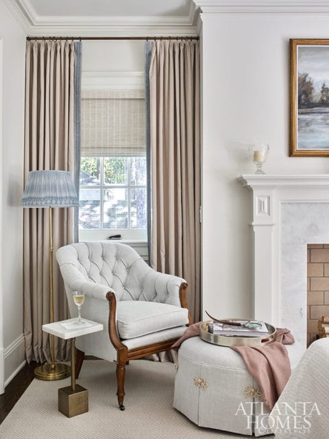 Light gray tufted armchair with wooden legs next to a window with beige curtains. A small marble-top side table with a brass base holds a glass of white wine.