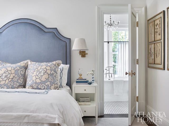 Bedroom with a light blue upholstered headboard and floral pillows, adjacent to a bathroom with a clawfoot tub and crystal chandelier.
