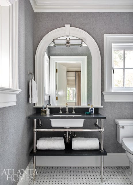 Gray textured wallpaper bathroom with arched mirror over a dark stone vanity with white sink, chrome fixtures, and two white towels on the shelf below.
