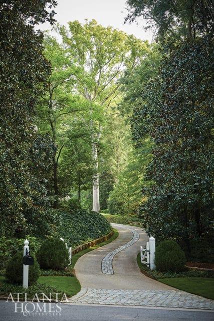 Curving driveway with cobblestone accents leading through a lush, green forest with mature trees and manicured hedges.