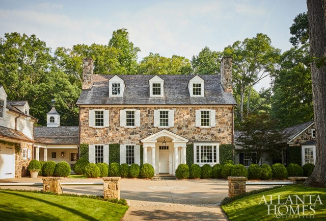 Stone-clad colonial home with white shutters and dormer windows, surrounded by manicured hedges and mature trees.