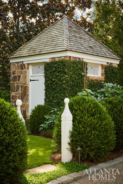 Stone and ivy-covered garden structure with a shingled hip roof, white door, and decorative window grilles, surrounded by manicured hedges and ornamental posts.