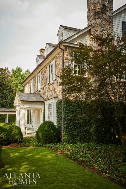 Stone and clapboard house with white shutters and a large stone chimney, surrounded by manicured hedges and trees.
