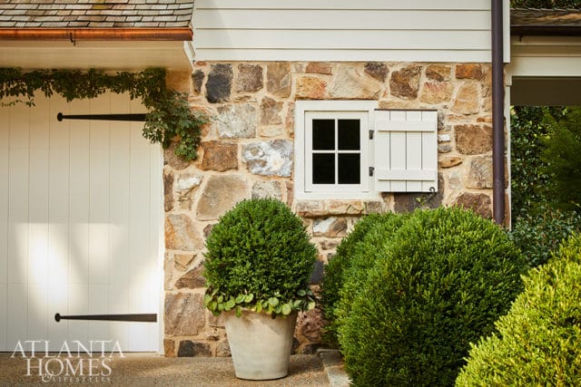 Stone facade with a white garage door, a small window with white shutters, and manicured boxwood bushes.
