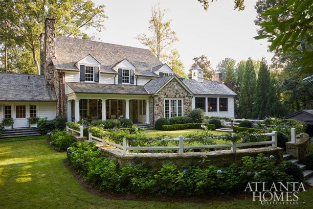 White clapboard house with stone accents, dormer windows, and a covered porch, surrounded by lush landscaping and a white picket fence.