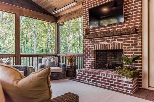 Screened porch with brick fireplace, mounted television, wicker seating, and wood-paneled ceiling overlooking a wooded area.