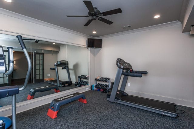 Home gym with treadmills, dumbbells, and a large mirror. The room has gray walls, a dark gray carpeted floor, and a black ceiling fan.