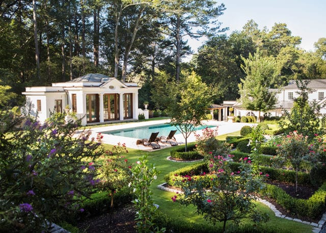 Formal garden with manicured hedges, flowering bushes, and a swimming pool with lounge chairs, adjacent to a white neoclassical pool house and main residence.