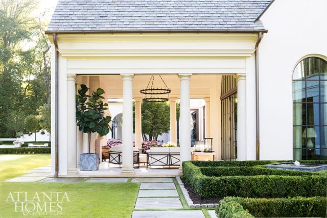 White stucco house with a covered patio supported by columns, featuring a large fiddle leaf fig tree, outdoor seating, and manicured hedges with a fountain.