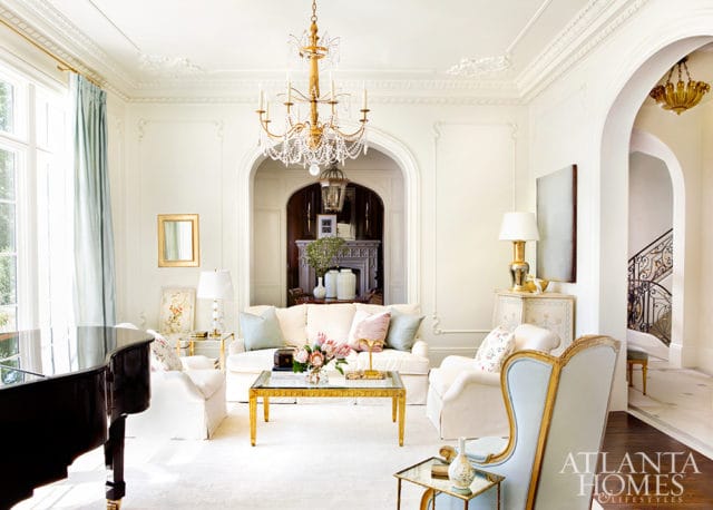 Elegant living room with a grand piano, white sofa, armchairs, and a crystal chandelier. Arched doorways reveal a fireplace and a staircase.