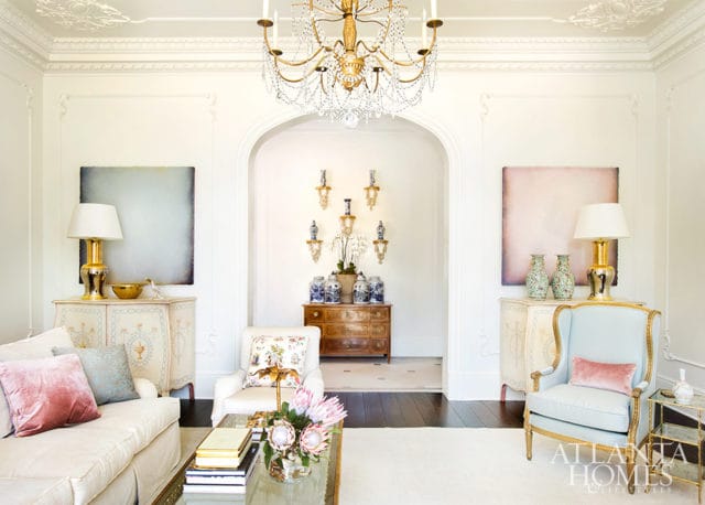 Living room with a white sofa, light blue armchair, ornate gold chandelier, and decorative chests. An arched doorway reveals a hallway with wall sconces and blue and white ginger jars.