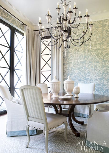 Round wooden dining table with upholstered chairs, ornate chandelier, and patterned wallpaper.