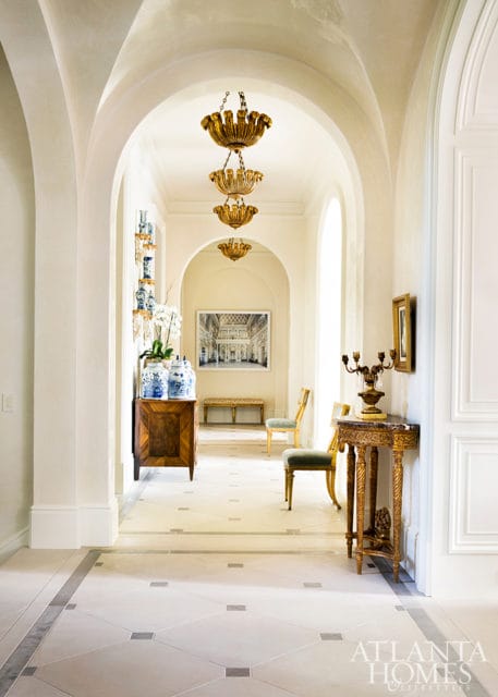 Hallway with arched ceilings, ornate gold chandeliers, marble floors with geometric inlay, antique wooden console, and blue and white porcelain vases.