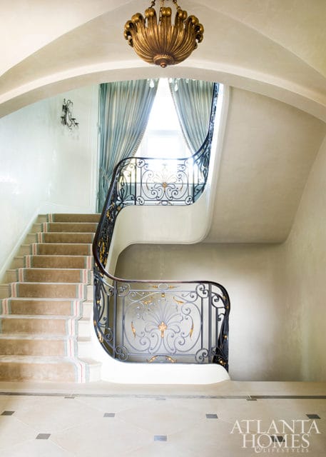 Curving staircase with ornate black and gold metal railing, plush carpet runner with red and blue stripes, and a dramatic gold chandelier hanging from an arched ceiling.