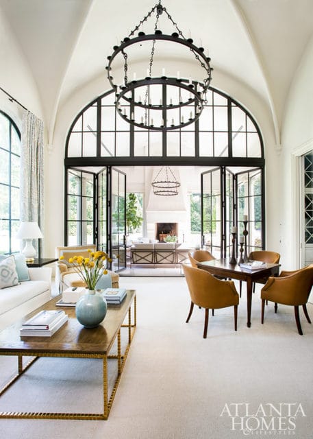 Living room with vaulted ceiling, large arched window with black grid, and two black chandeliers. White sofa, gold coffee table, and four tan armchairs around a dark wood table.