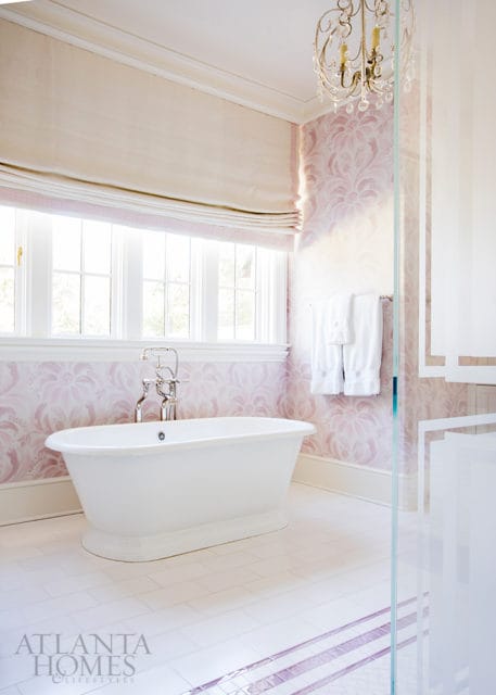 White freestanding bathtub in a bathroom with pink floral wallpaper, white tile floor, and a crystal chandelier.