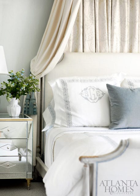 Bedroom with a white upholstered headboard, draped canopy, and white bedding with blue embroidered trim and a monogrammed pillow. A mirrored nightstand holds a vase of flowers.
