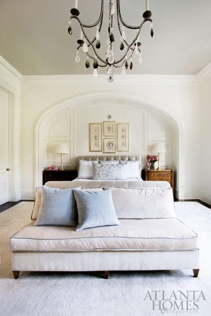 Cream upholstered sofa with light blue pillows in front of a bed with a tufted headboard, flanked by dark wood nightstands. A crystal chandelier hangs from the ceiling.