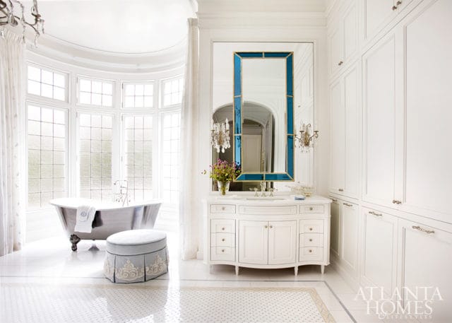 White bathroom with freestanding tub, ornate vanity, large blue-framed mirror, and floor-to-ceiling white cabinetry.