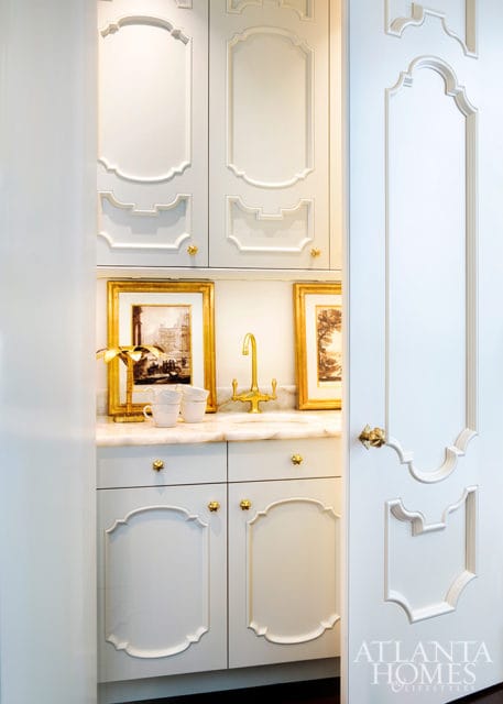 White built-in cabinetry with ornate molding and gold hardware, featuring a marble countertop, gold faucet, and framed artwork above a sink.