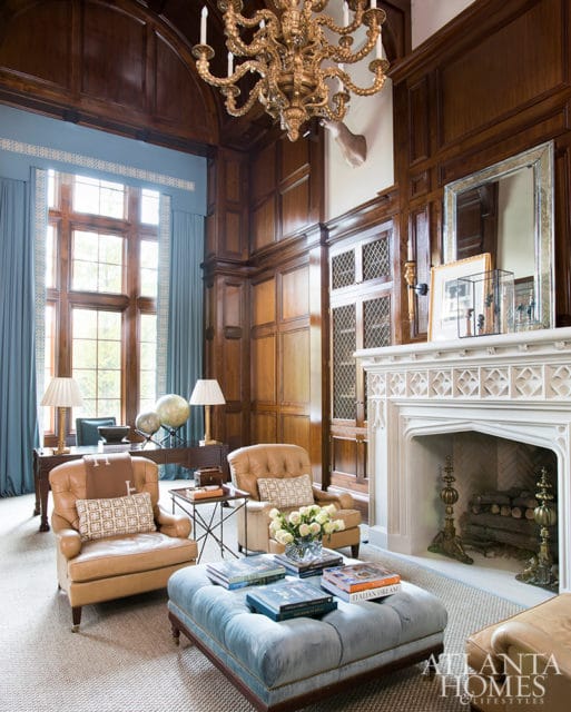 Library with dark wood paneling, a large stone fireplace, tan leather armchairs, and a light blue tufted ottoman topped with books.