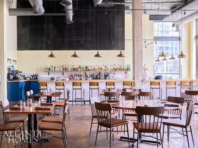 Restaurant interior with a long bar stocked with bottles, white subway tile backsplash, and wooden stools. Dining tables with brown leather chairs are set with place settings. Exposed ductwork and industrial pendant lights hang from the ceiling.