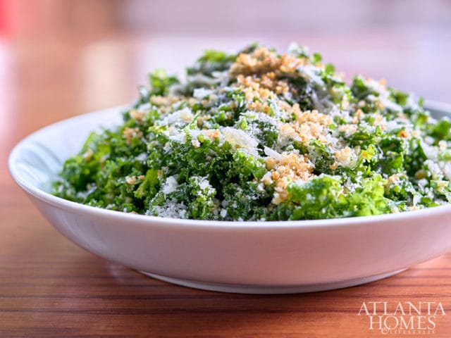 Close-up of a white bowl filled with chopped kale salad topped with shredded cheese and breadcrumbs, set on a wooden table.