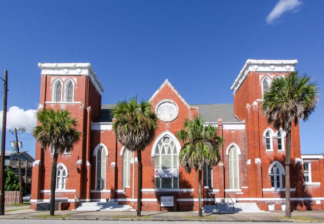 Red brick church with white trim, arched windows, and two towers, surrounded by palm trees under a bright blue sky.