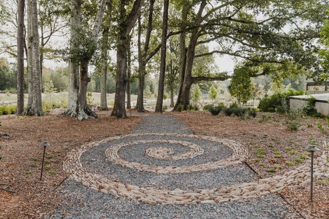 Gravel pathway with a spiral design made of light-colored stones, surrounded by trees and landscaping.