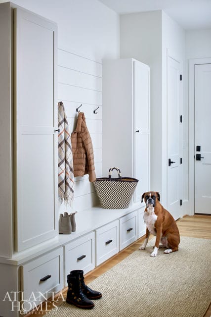 White mudroom with shiplap wall, built-in bench with drawers, tall cabinets, and a boxer dog sitting on a woven rug.