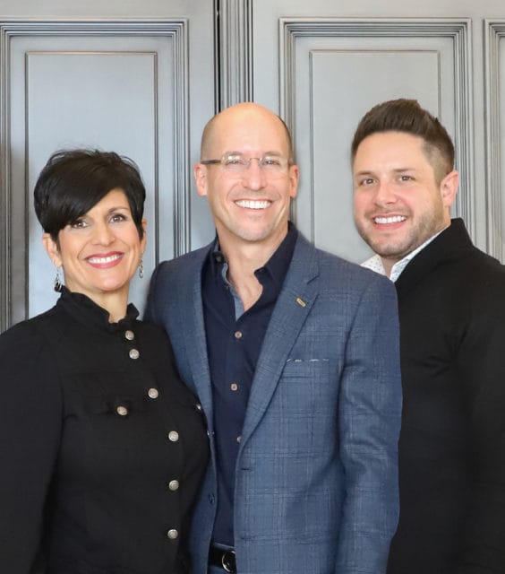 Three people smiling, two men and one woman, standing in front of a grey paneled wall.