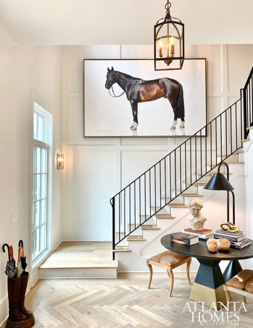 Foyer with herringbone wood floors, a large painting of a brown horse, a black metal staircase railing, and a round table with decorative objects and books.