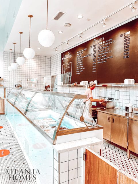 Ice cream shop interior with a glass display case filled with ice cream, a large chalkboard menu listing flavors, and a person in an apron behind the counter.