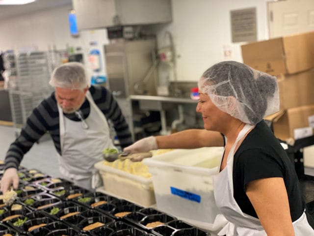 Two volunteers wearing hairnets and aprons scoop food into black takeout containers in a commercial kitchen.