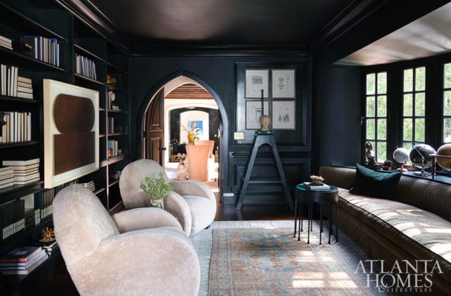 Dark moody library with floor-to-ceiling bookshelves, plush cream armchairs, a curved brown sofa, and an arched doorway leading to another room.